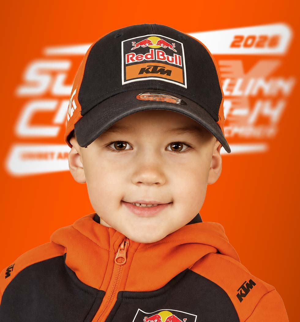 Smiling young boy in an orange and black KTM jacket and Red Bull cap against a bright orange branded backdrop.